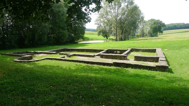 Die Umrisse eines alten Römischen Gebäudes im Boden einer Wiese zu sehen. Drum herum befinden sich Bäume und ein Weiher im Hintergrund.