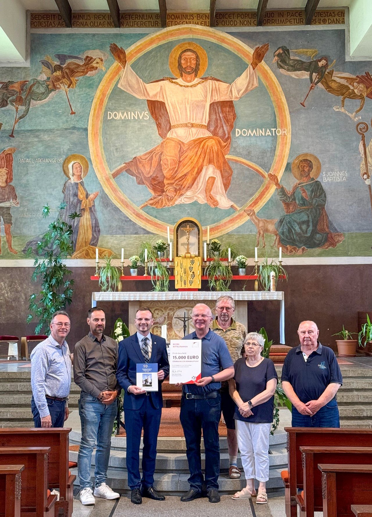 Bezirkstagspräsident Peter Daniel Forster steht zusammen mit den beteiligten Personen im sanierten Gebäude mit dem Förderbescheid in der Hand.