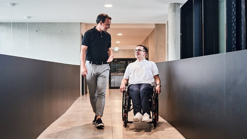 Zwei Menschen bewegen sich auf die Kamera zu und unterhalten sich dabei. Ein Mann läuft, der andere fährt im Rollstuhl.