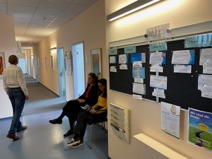 Szene im Flur des Sozialpsychiatrischen Diensts Fürth. Drei Personen warten. Im Vordergrund hängt eine Pinnwand, die die aktuellen Gruppenangebote abbildet.