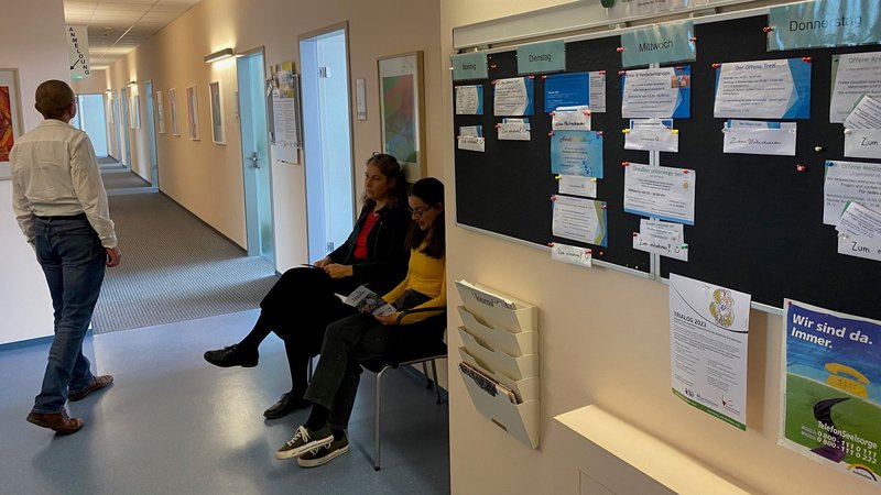 Szene im Flur des Sozialpsychiatrischen Diensts Fürth. Drei Personen warten. Im Vordergrund hängt eine Pinnwand, die die aktuellen Gruppenangebote abbildet.