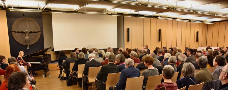 Ein voll gefüllter Saal. Das Publikum sitzt auf Stühlen und wartet, dass es auf der Bühne losgeht.