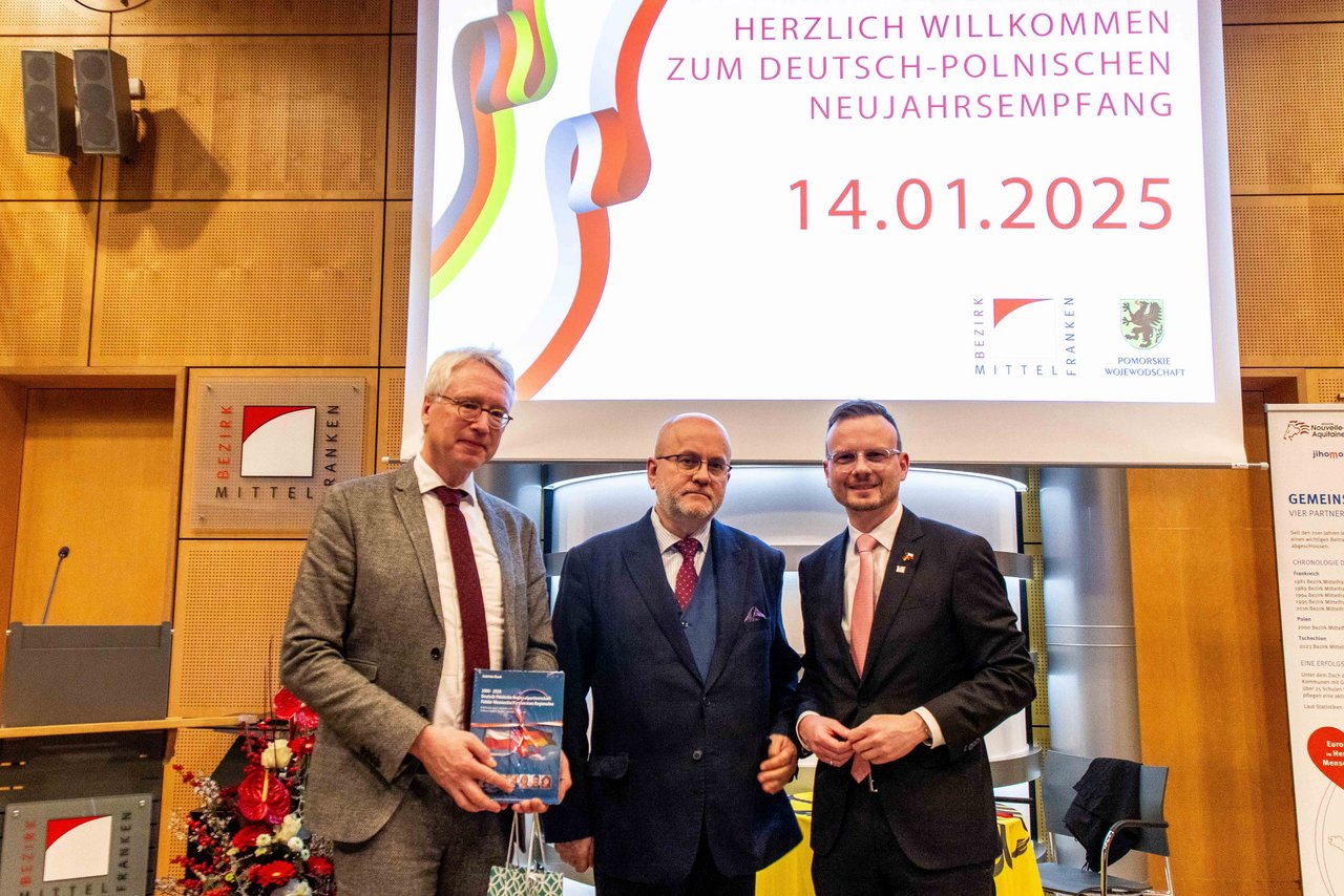 Prof. Dr. Peter Oliver Loew, Rafał Wolski und Bezirkstagspräsident Peter Daniel Forster stehen beim deutsch-polnischen Neujahrsempfang für ein Gruppenfoto vor der Leinwand im Sitzungssaal des Bezirksrathauses in Ansbach.