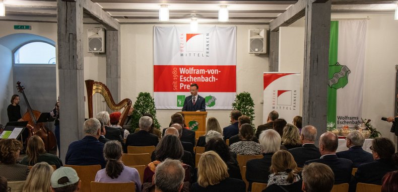Bezirkstagspräsident Peter Daniel Forster steht in einem Saal vorne auf dem Podium und spricht zum Publikum, das auf Stühlen vor ihm sitzt.