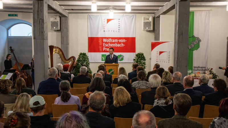 Bezirkstagspräsident Peter Daniel Forster steht in einem Saal vorne auf dem Podium und spricht zum Publikum, das auf Stühlen vor ihm sitzt.