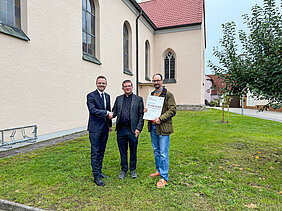 Drei Männer halten Förderbescheid in den Händen vor einer Kirche