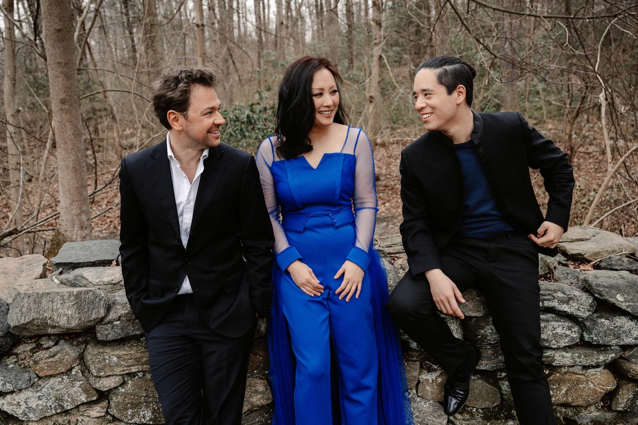 Die drei Mitglieder des Sitkovetsky Trios stehen im Wald an eine kleine Steinmauer angelehnt und lachen sich an.