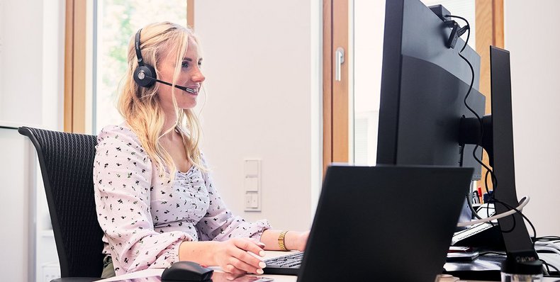 Eine Dame sitzt an ihrem Schreibtisch am PC, schaut auf ihren Bildschirm, hat ein Headset auf und telefoniert gerade.