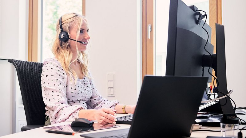 Eine Dame sitzt an ihrem Schreibtisch am PC, schaut auf ihren Bildschirm, hat ein Headset auf und telefoniert gerade.