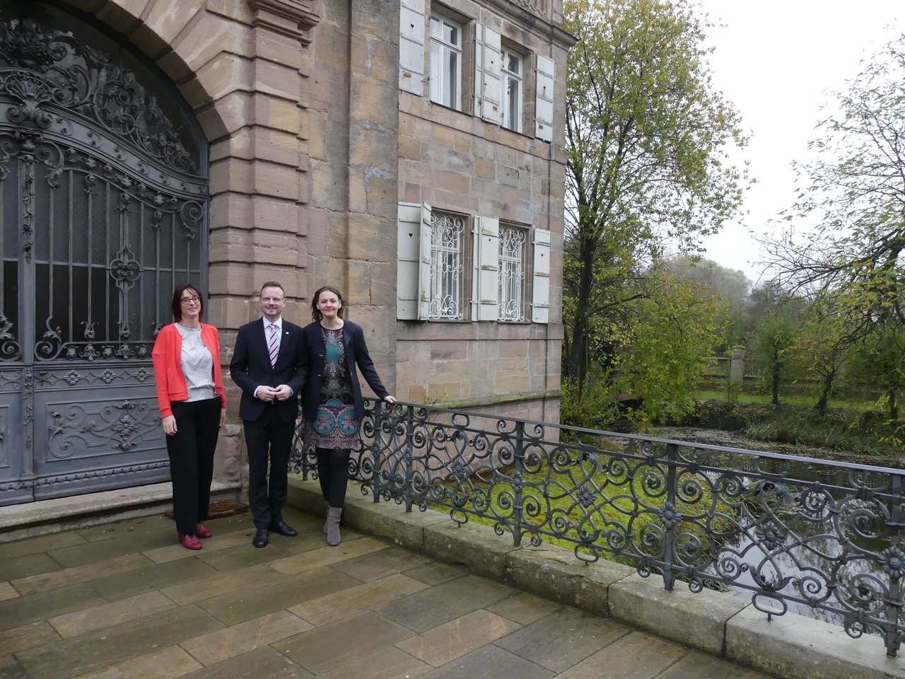 Kulturreferentin und Bezirksheimatpflegerin Dr. Annett Haberlah-Pohl, Bezirkstagspräsident Peter Daniel Forster und die Künstlerische Leiterin Franziska Hölscher stehen auf der Eingangsbrücke des Schlosses Dürrenmungenau