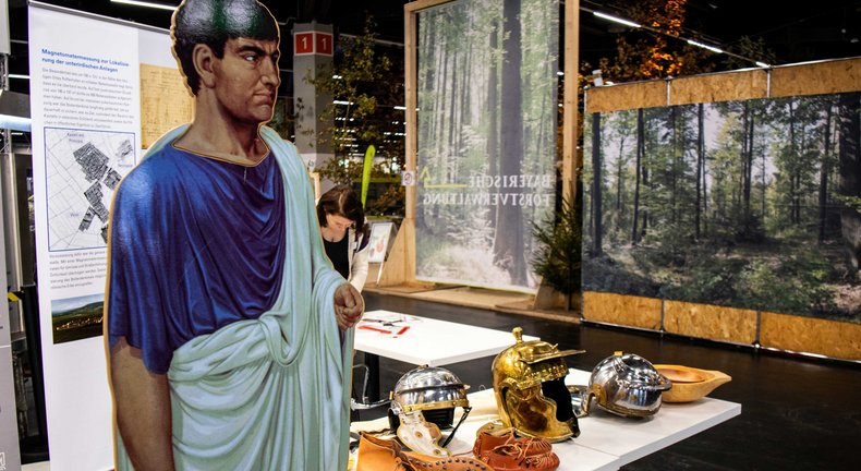 Ein auf Holz aufgemalter und in Lebensgröße aufgestellter Römer, der neben einem Tisch steht, auf dem Utensilien aus der Römerzeit ausgestellt sind.