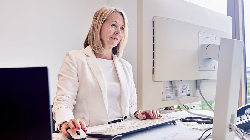 Eine Frau steht an ihrem Schreibtisch und schaut auf ihren PC-Bildschirm. Die eine Hand an der Maus, die andere Hand an der Tastatur.