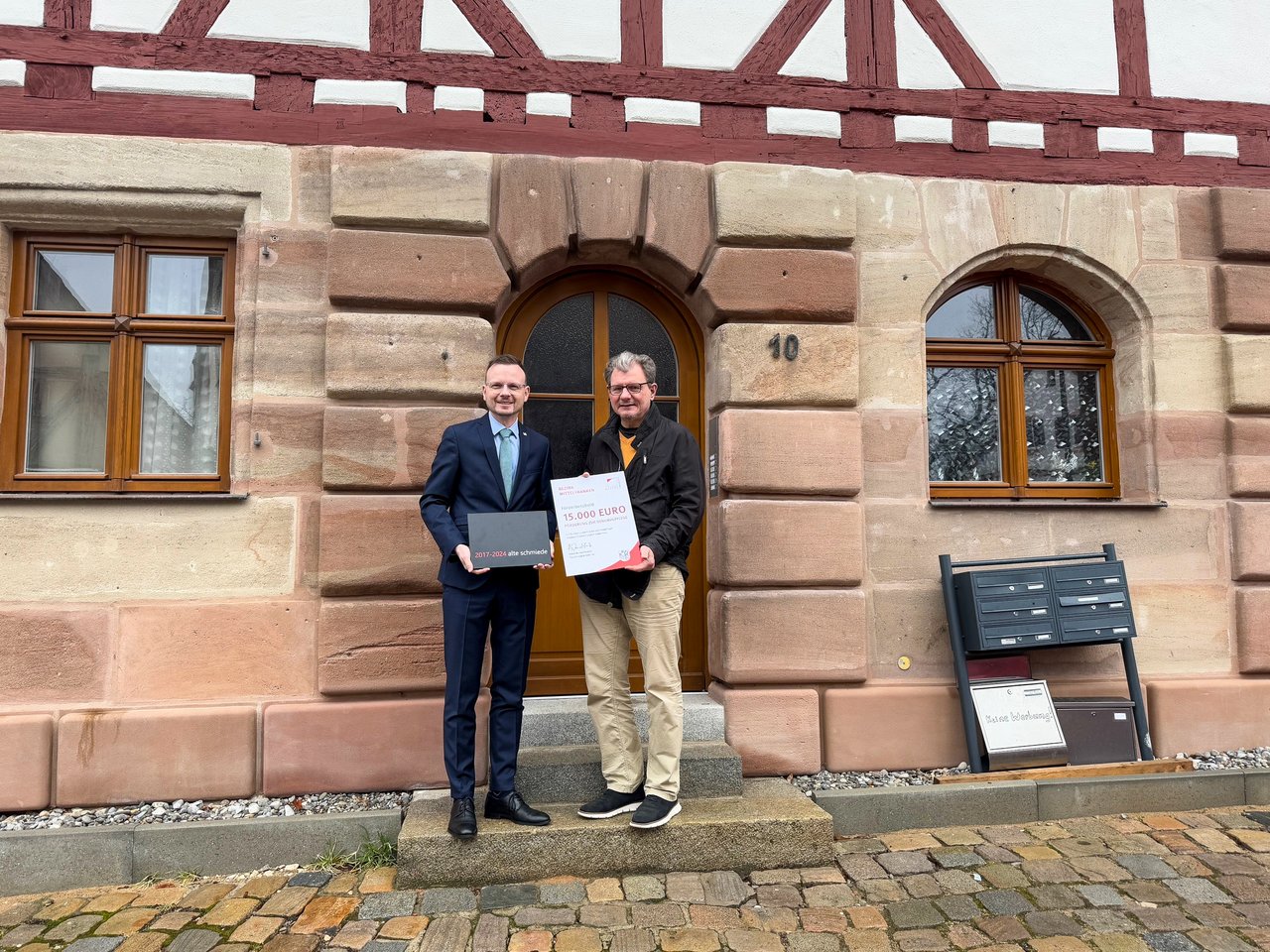 Bezirkstagspräsident Peter Daniel Forster überreicht den Förderbescheid in Nürnberg an Stefan Guertler. Beide stehen vor dem sanierten Gebäude.