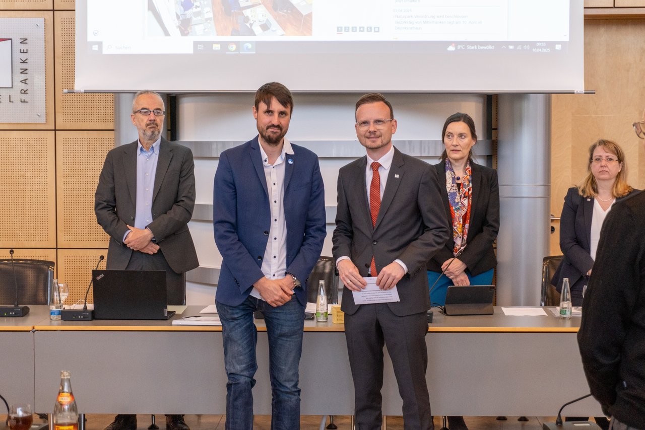 Titus Schüller und Bezirkstagspräsident Peter Daniel Forster stehen im Sitzungssaal des Bezirksrathauses vor dem Plenum und posieren für ein Foto. Im Hintergrund stehen Fritz Weispfenning, Christa Heckel und Christina Lowig.
