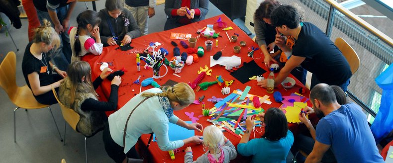 Blick von oben auf einen großen Tisch, auf dem gebastelt wird. Es stehen zahlreiche Kinder um den Tisch herum.