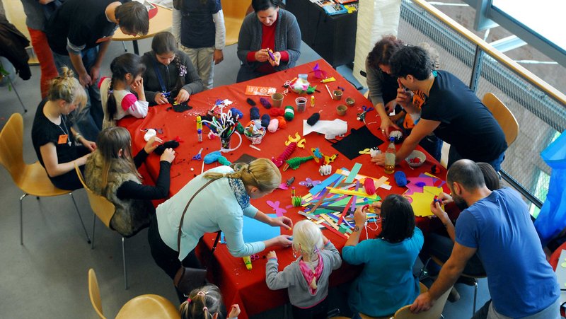 Blick von oben auf einen großen Tisch, auf dem gebastelt wird. Es stehen zahlreiche Kinder um den Tisch herum.