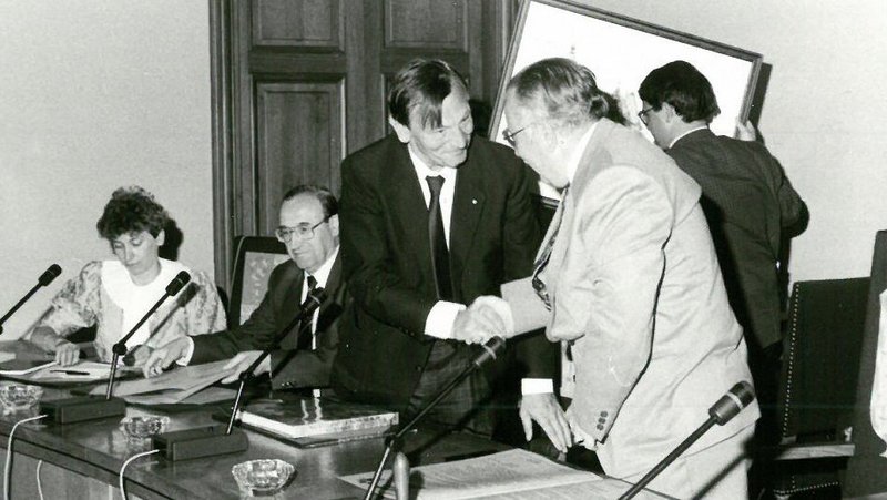 Zwei Menschen stehen an einem Rednerpult und geben sich gerade die Hand, bevor sie sich setzen. Das Foto ist sehr alt und in schwarz/weiß aufgenommen.