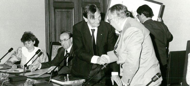 Zwei Menschen stehen an einem Rednerpult und geben sich gerade die Hand, bevor sie sich setzen. Das Foto ist sehr alt und in schwarz/weiß aufgenommen.