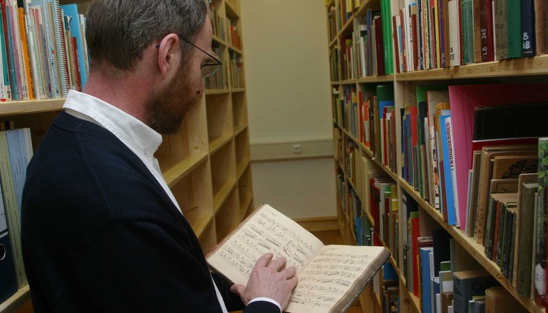 Ein Mann steht in einer Bibiliothek und schaut gerade in ein Buch rein. Man schaut ihm über die Schulter.