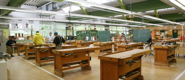 Ein Blick in eine Holzwerkstatt. Es stehen zahlreiche Holzarbeitstische in einem großen Arbeitsraum.