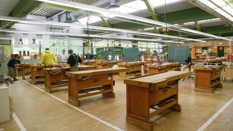 Ein Blick in eine Holzwerkstatt. Es stehen zahlreiche Holzarbeitstische in einem großen Arbeitsraum.