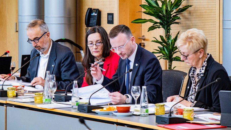 Bezirkstagspräsident Peter Daniel Forster sitzt mit weiteren Personen vorne am Pult des Sitzungssaals und leitet eine Sitzung. Er greift gerade an sein Mikrofon, um etwas hineinzusprechen.
