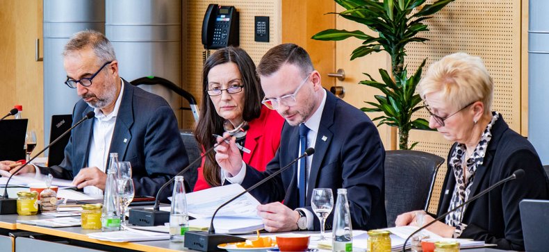 Bezirkstagspräsident Peter Daniel Forster sitzt mit weiteren Personen vorne am Pult des Sitzungssaals und leitet eine Sitzung. Er greift gerade an sein Mikrofon, um etwas hineinzusprechen.