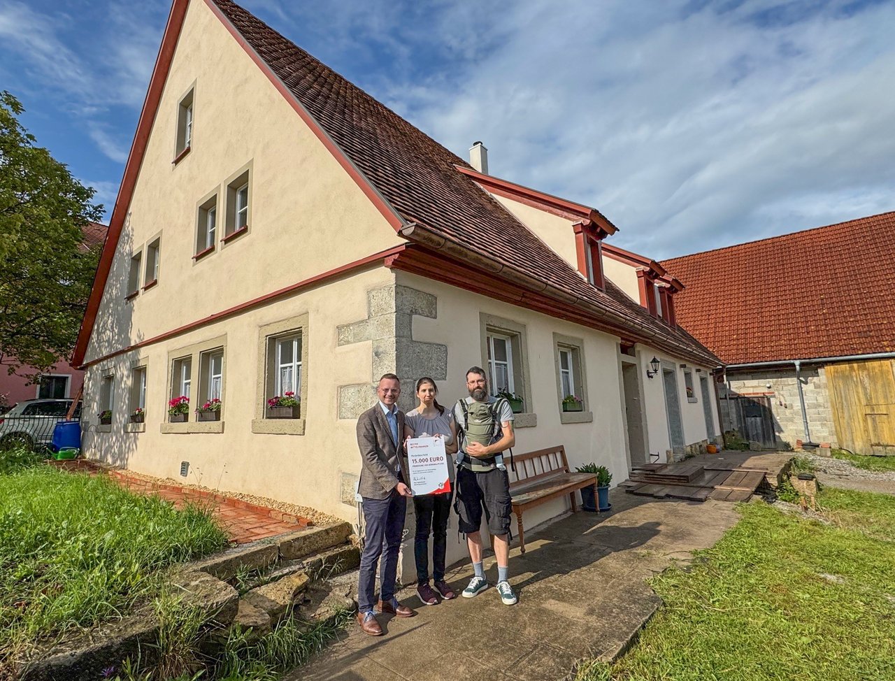 Bezirkstagspräsident Peter Daniel Forster steht zusammen mit den beteiligten Personen vor dem sanierten Gebäude mit dem Förderbescheid in der Hand.