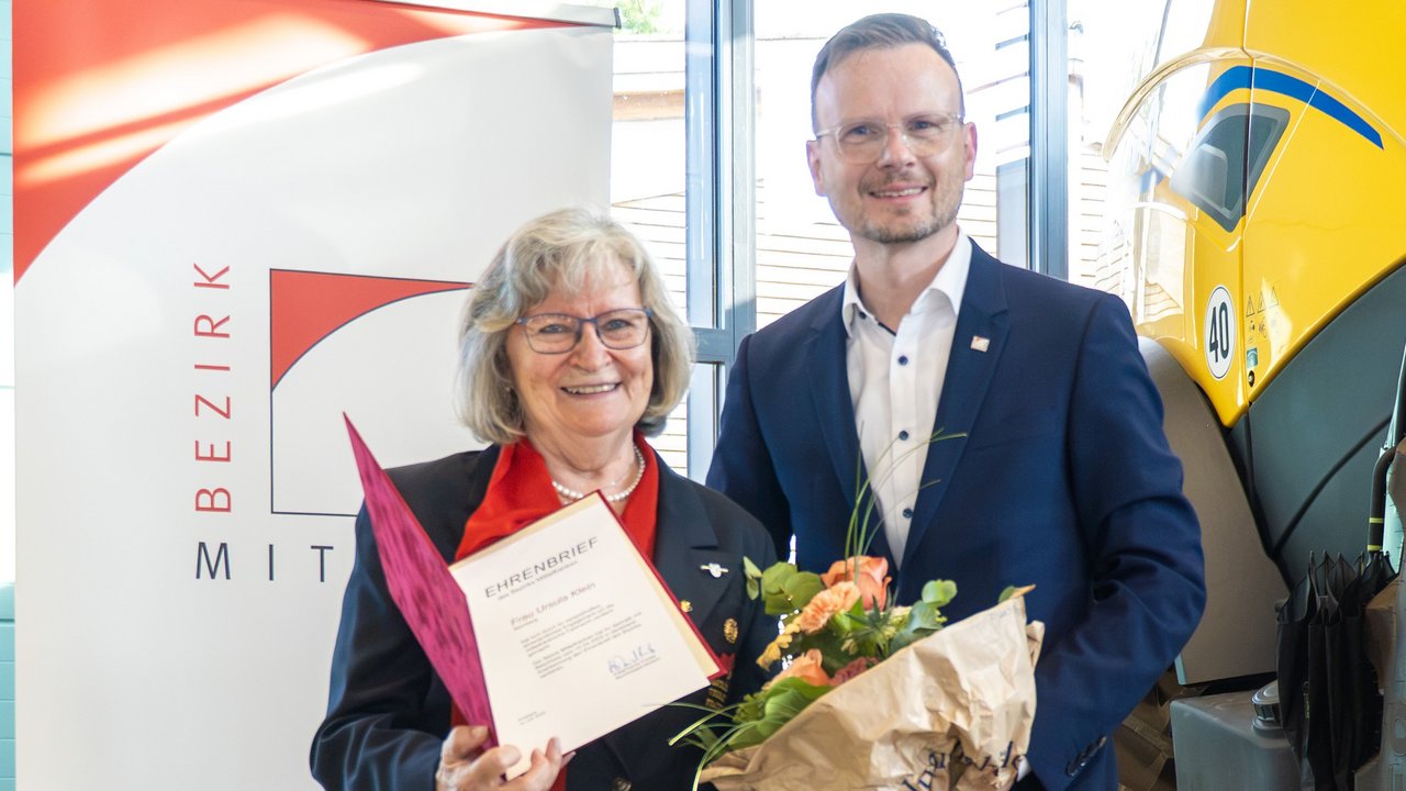 Gruppenbild der beiden aufgezählten Personen. Ursula Klein hält dabei den verliehenen Ehrenbrief in die Kamera, Bezirkstagspräsident Peter Daniel Forster einen Blumenstrauß. Die beiden stehen vor einem Rollup des Bezirks Mittelfranken.