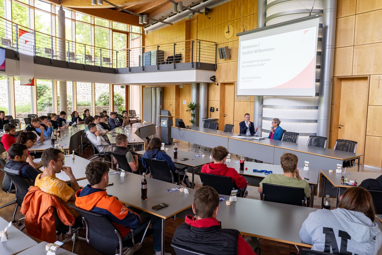 Bezirkstagspräsident Peter Daniel Forster sitzt vorne am Pult im Sitzungssaal des Bezirksrathauses und gibt den französischen Austauschschülern einen Kurzüberblick über den Bezirk Mittelfranken.