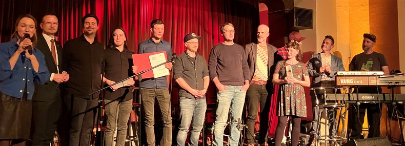 Die Preisträger des Pop Preises 2024 stehen zusammen mit Bezirkstagspräsident Peter Daniel Forster und der Moderatorin nebeneinander auf einer Bühne.