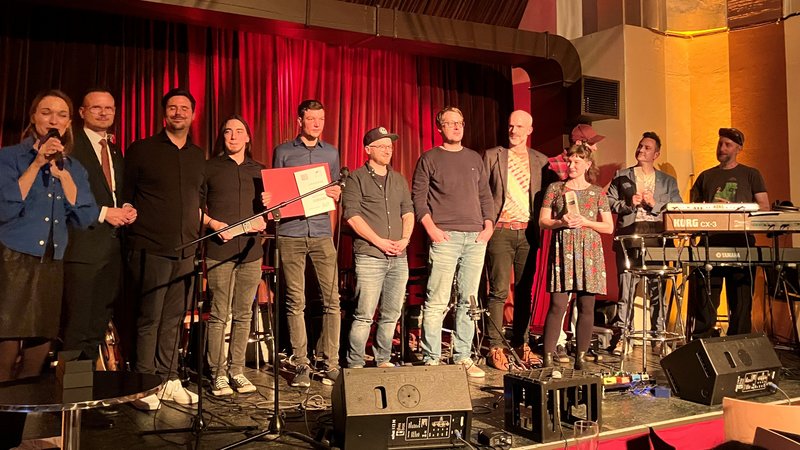 Die Preisträger des Pop Preises 2024 stehen zusammen mit Bezirkstagspräsident Peter Daniel Forster und der Moderatorin nebeneinander auf einer Bühne.