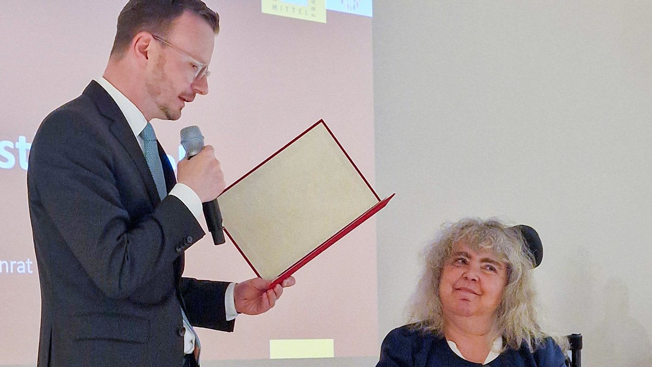 Bezirkstagspräsident Peter Daniel Forster hat den Ehrenbrief in der Hand und liest die Urkunde vor. Neben ihm sitzt Angelika Feisthammel.