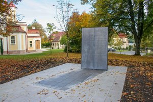 Bild des Euthanasie-Mahnmals auf dem Gelände der Bezirkskliniken in Ansbach. Eine metallene Gedenktafel mit dem Namen der Opfer steht auf einem asphaltierten Fundament. Im Hintergrund sind Bäume und Gebäude der Bezirkskliniken zu sehen.