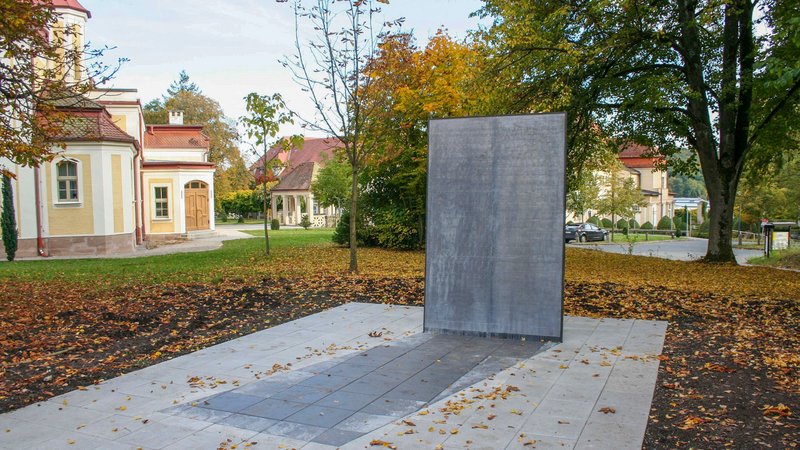 Bild des Euthanasie-Mahnmals auf dem Gelände der Bezirkskliniken in Ansbach. Eine metallene Gedenktafel mit dem Namen der Opfer steht auf einem asphaltierten Fundament. Im Hintergrund sind Bäume und Gebäude der Bezirkskliniken zu sehen.