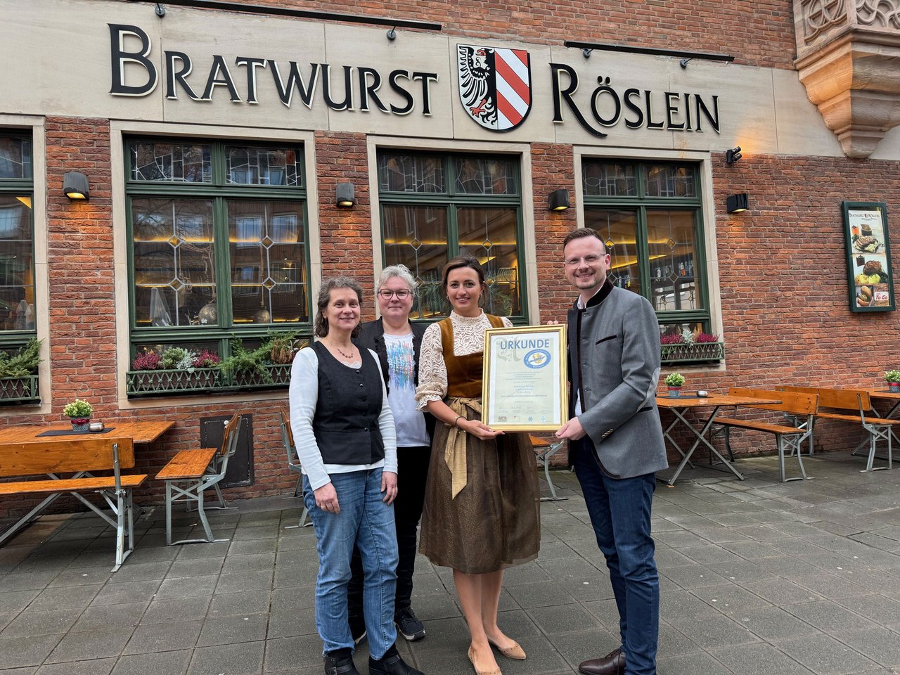 Die genannten Personen bei der nachgeholten Ehrung in Nürnberg stehen vor dem Gebäude des Wirtshaus Bratwurst Röslein. Die Wirtin und Bezirkstagspräsident Forster halten die Urkunde in die Kamera.