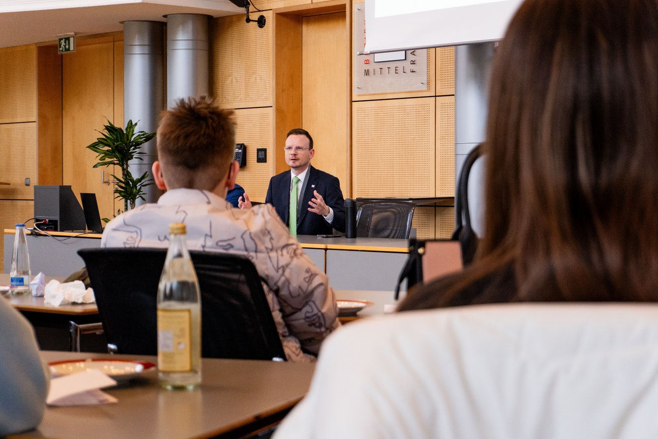 Blick über die Schultern der Schüler hinweg nach vorne ans Rednerpult, an dem Bezirkstagspräsident Peter Daniel Forster sitzt und zu den Schülern spricht.