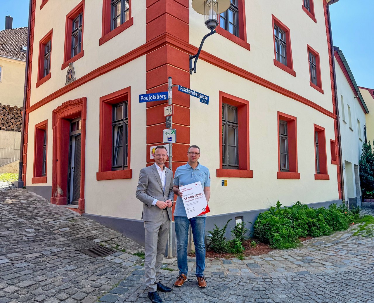 Bezirkstagspräsident Peter Daniel Forster steht zusammen mit den beteiligten Personen vor dem sanierten Gebäude mit dem Förderbescheid in der Hand.