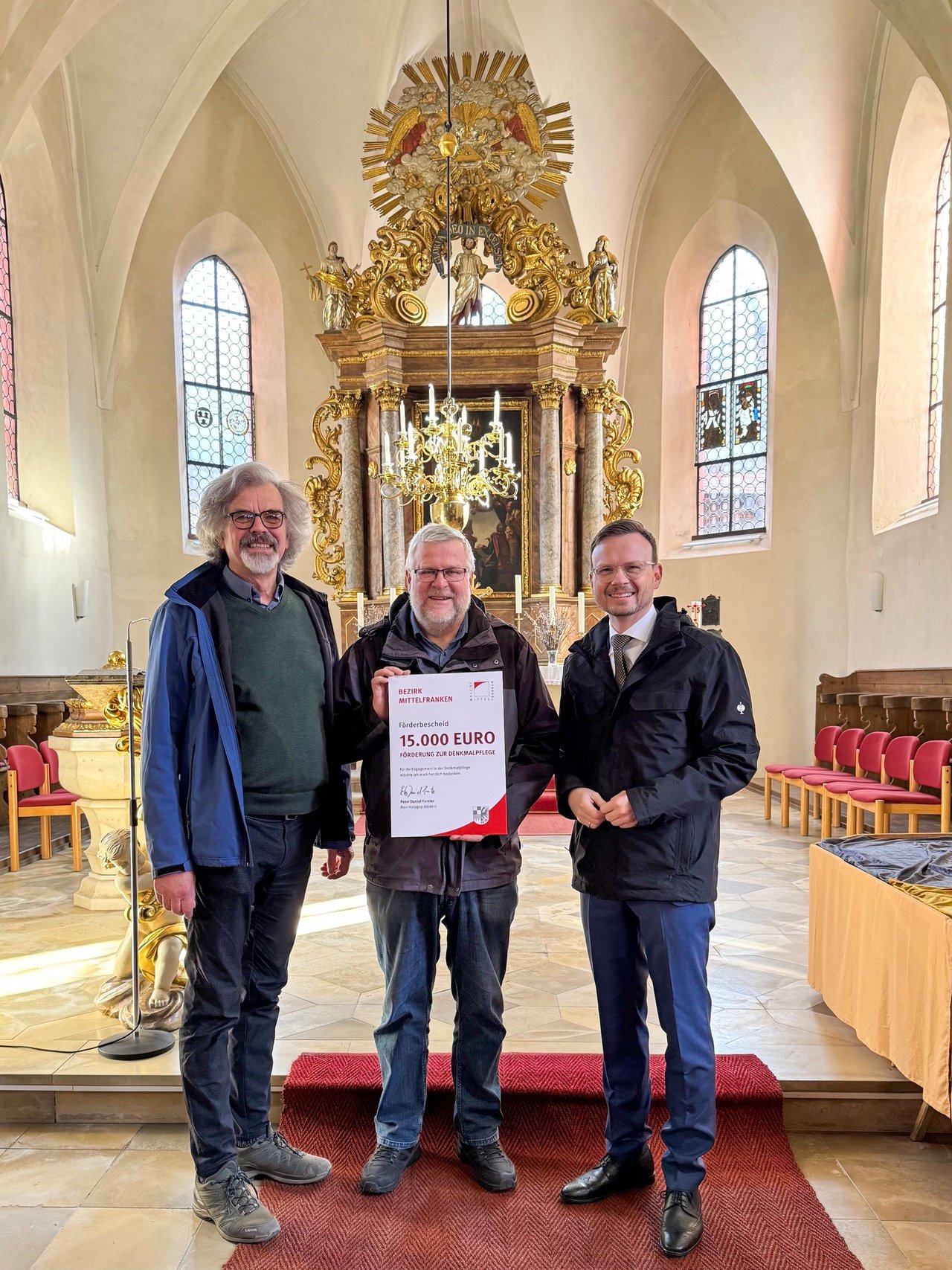 Die drei beteiligten Personen stehen in der Kirche vor dem Altar und halten den Förderbescheid hoch.