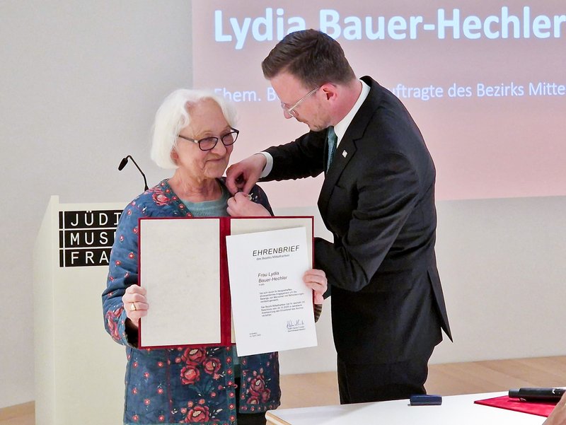 Bezirkstagspräsident Peter Daniel Forster steckt Lydia Bauer-Hechler die dazugehörige Anstecknadel des Ehrenbriefs an, während Bauer-Hechler die Urkunde in der Hand hält.
