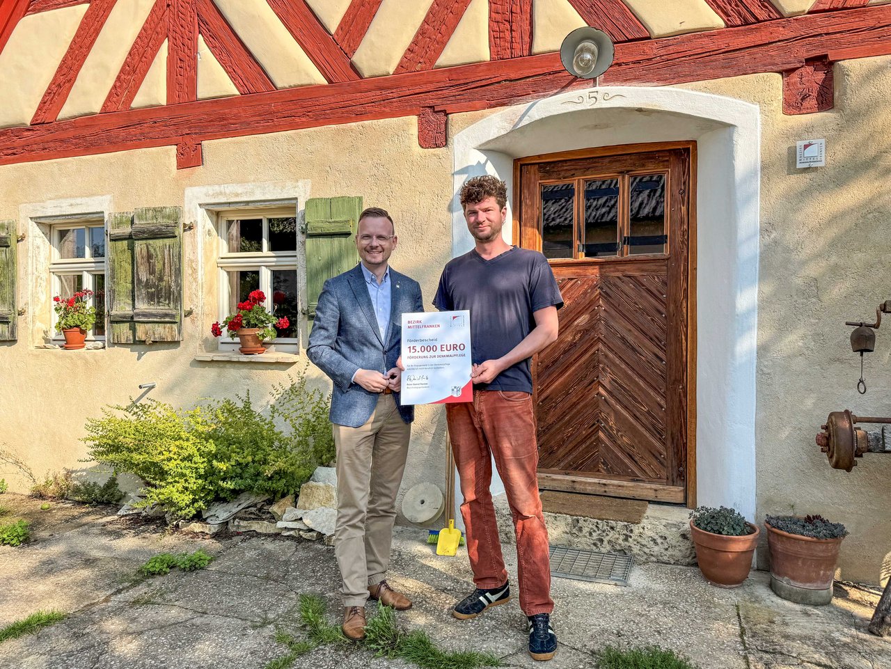 Zwei Männer stehen vor Fachwerkhaus und halten einen Scheck