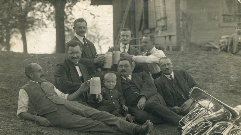 Archivbild von 1921: Musikanten einer Kapelle liegen auf einer Wiese, lachen in die Kamera und halten Bierkrüge in der Hand.