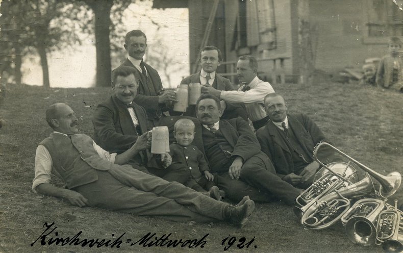 Archivbild von 1921: Musikanten einer Kapelle liegen auf einer Wiese, lachen in die Kamera und halten Bierkrüge in der Hand.
